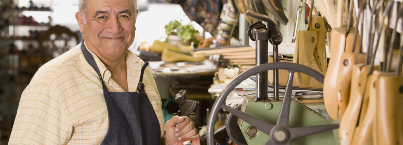 elderly shoe cobbler gentleman smiling with one hand on shoe mending machine, shoe trees, shoes and equipment in background