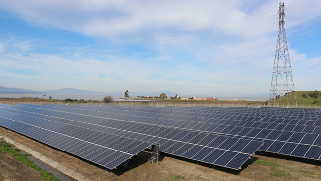 large scale solar system on dirt ground, electrical tower, clear sky, clouds, mountains in distance