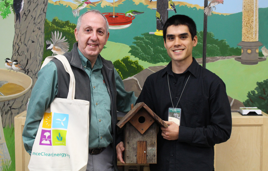 two staff members hold tote bag and wooden bird house, both smile in front of painted bird mural