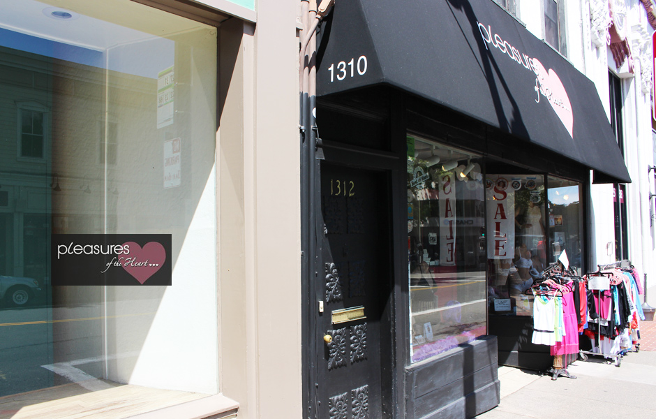 outside store front, signage says Pleasure of the Heart, shows pink heart, sidewalk sale rack of clothing, window says sale