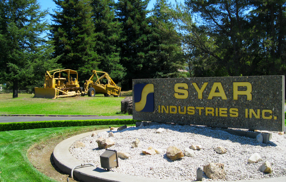 grass lawn, tall pine trees, official sign, says Syar Industrics Inc., nonoperational construction vehicles in background