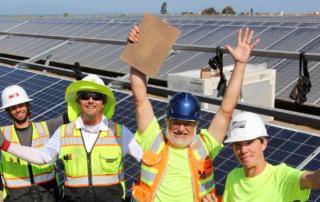 four solar workers with construction attire, raise hands in excitement in front of commercial solar project, clear sky