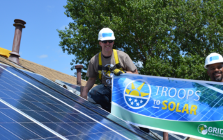 two solar installers next to solar system on rooftop of home, holding banner, says Troops to Solar, GRID Alternatives