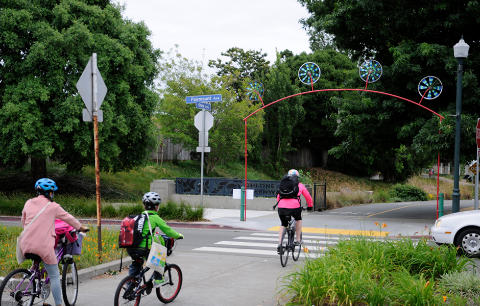 City of El Cerrito bike path crossing Fairmount Avenue, three bicyclists with helmets, car stopped, lush trees, overcast sky