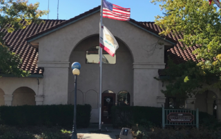 front entrance of Calistoga City Hall, American and California States flags blowing on pole, bushes, trees