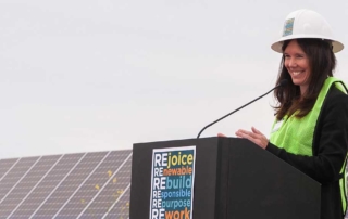 MCE Chief Executive Officer, Dawn Weisz with hard hat, safety vest, speaks in microphone on podium, solar panels behind her