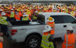 over one hundred solar workers, hard hats, construction vests, wait for briefing, solar panels, several pick up trucks