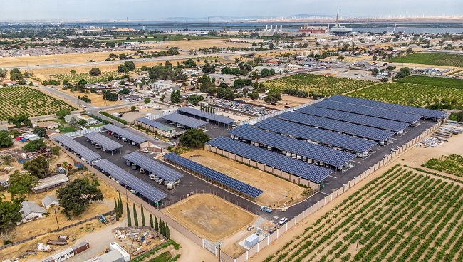 Drohnenansicht eines großen Sonnensystems, das sich auf mehreren Carport-Schattenstrukturen, landwirtschaftlichen Flächen und Delta im Hintergrund ausbreitet