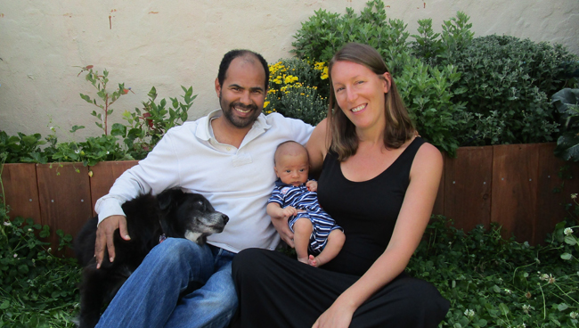 happy father with dog in his lap close to smiling mother holding new born baby in right hand, sitting in backyard garden