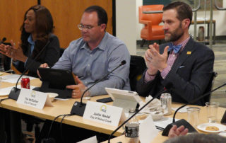 MCE Board Members, Sue Higgins, Shanelle Scales-Preston, and Justin Wedel sit and clap, Tim McGallian sits, looks at Ipad