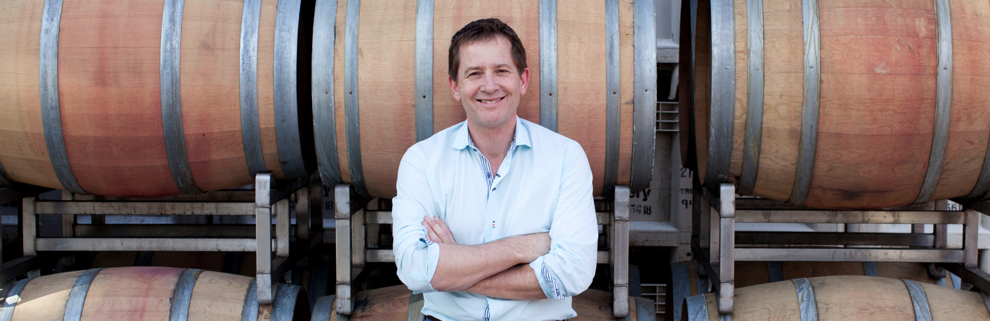 Michael Honig, owner of Honig Vineyard and Winery, smiles, crosses arms in front of six wine barrels inside cellar