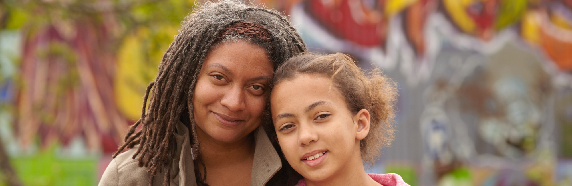 mother and daughter duo smile pleasantly in front of out of focus mural background in urban area